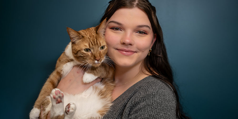 A woman with long dark hair smiles while holding an orange and white cat in her arms. The background is a solid dark teal color.