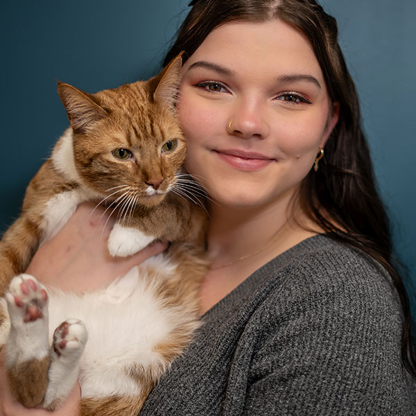 A person with long dark hair smiles while holding an orange and white cat against a teal background. The cat has green eyes and relaxed paws. The person is wearing a gray sweater and has a nose ring.