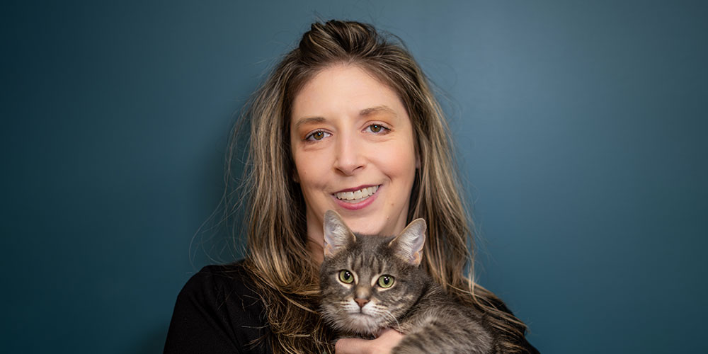 A woman with long brown hair smiling and holding a gray tabby cat. She is standing against a solid dark blue background. The cat has green eyes and is looking at the camera.