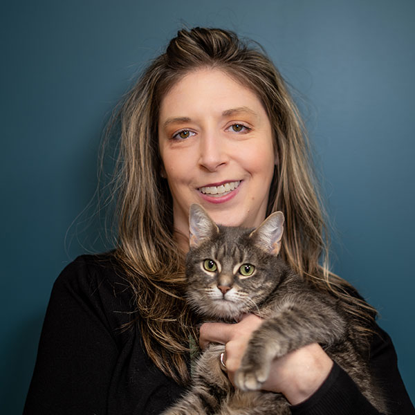 A woman with long hair smiles while holding a gray tabby cat in her arms. The background is a solid dark teal color.