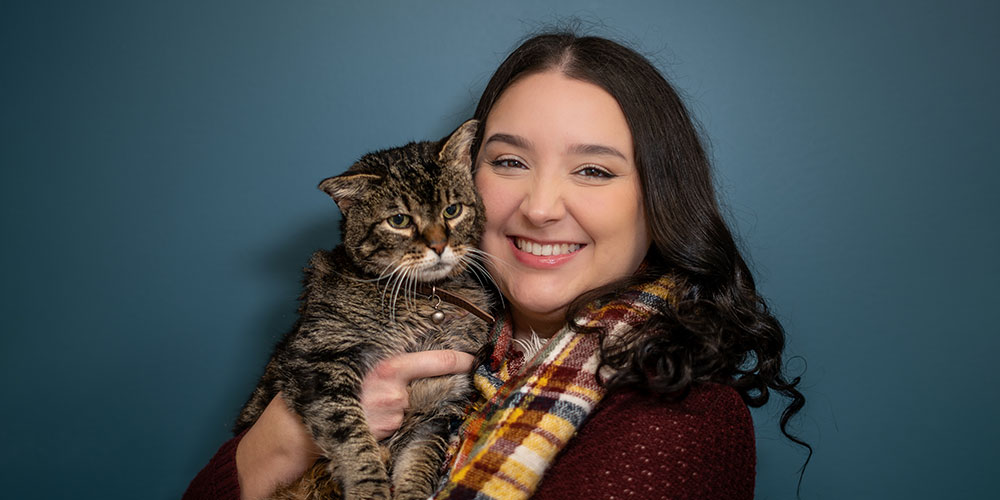 A woman with long dark hair smiles while holding a tabby cat. She wears a colorful plaid scarf and a maroon sweater. The background is teal, adding contrast to the warm colors of her outfit and the cat's fur.