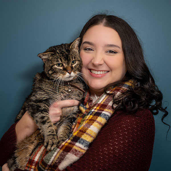 A woman with long dark hair smiles while holding a tabby cat. She is wearing a colorful plaid scarf and a maroon sweater. The background is a solid blue color.