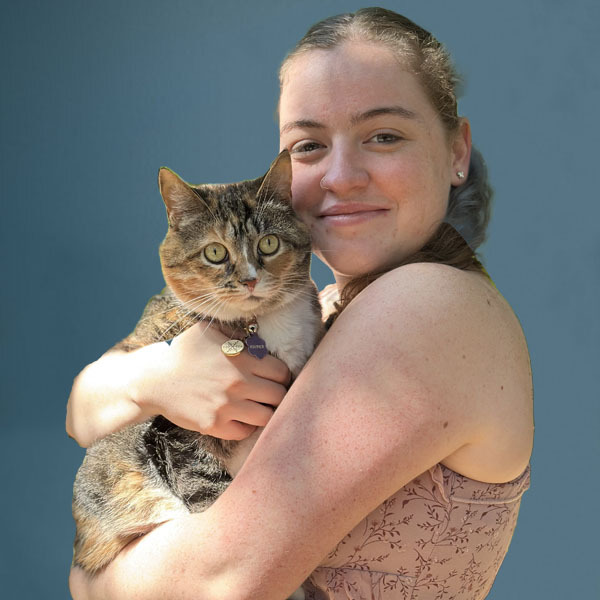 A woman smiling and holding a brown and white tabby cat in her arms, standing against a plain blue background. The cat looks at the camera with wide eyes.