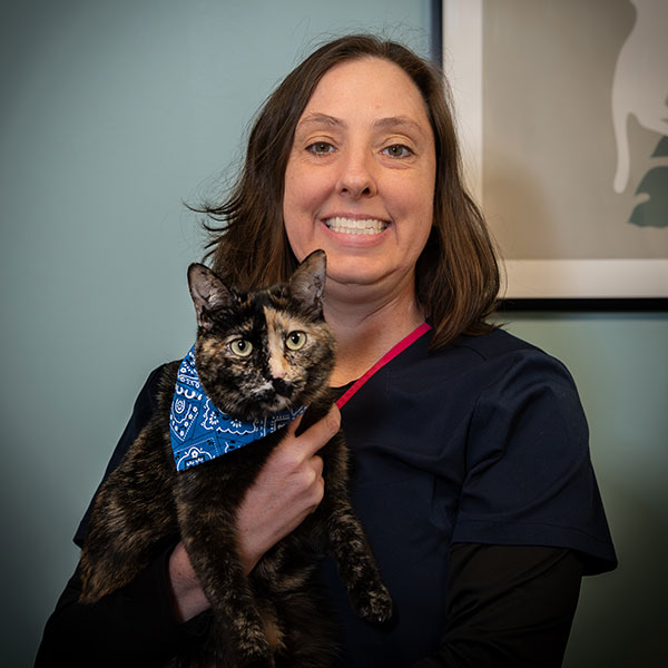 A woman with shoulder-length brown hair, wearing dark scrubs, smiles while holding a tortoiseshell cat dressed in a blue bandana, standing indoors next to a framed picture on a light blue wall.