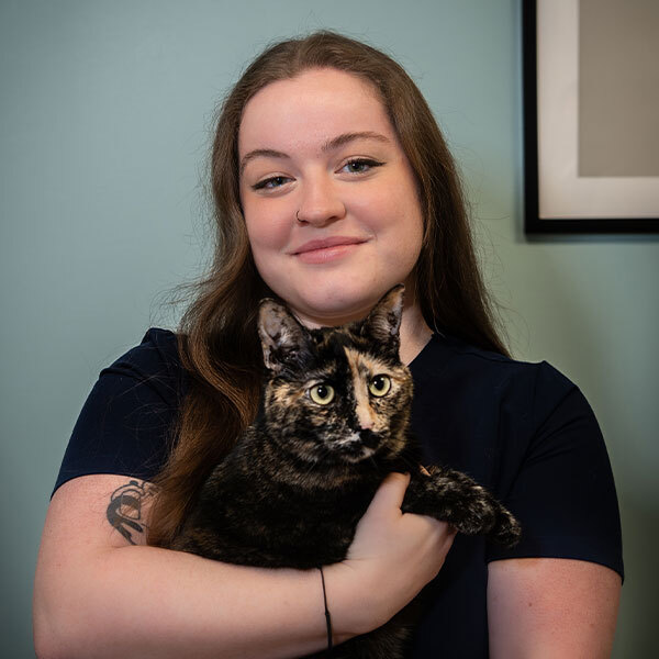 A woman with long brown hair smiles while holding a tortoiseshell cat in her arms, standing against a light blue wall with part of a framed picture visible in the background.