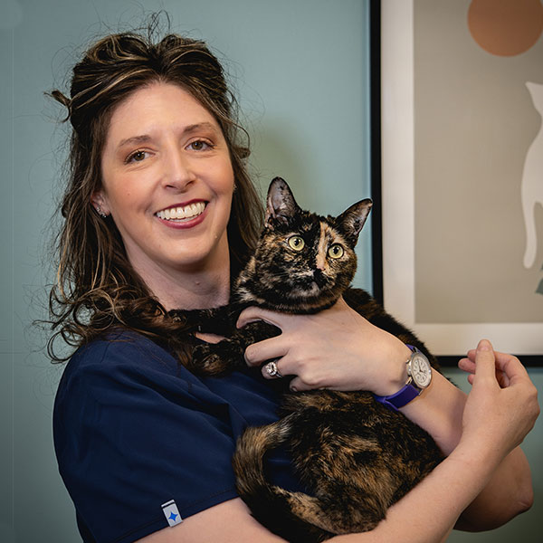A woman in navy scrubs smiles while holding a black and orange tortoiseshell cat in her arms, standing indoors next to a framed cat-themed artwork on a blue wall.